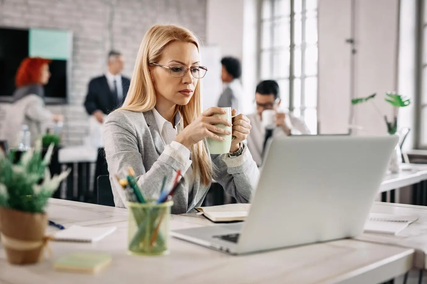 A beautiful, pensive businesswoman sits at her office desk, drinking tea and reading something on her laptop, with people in the background.