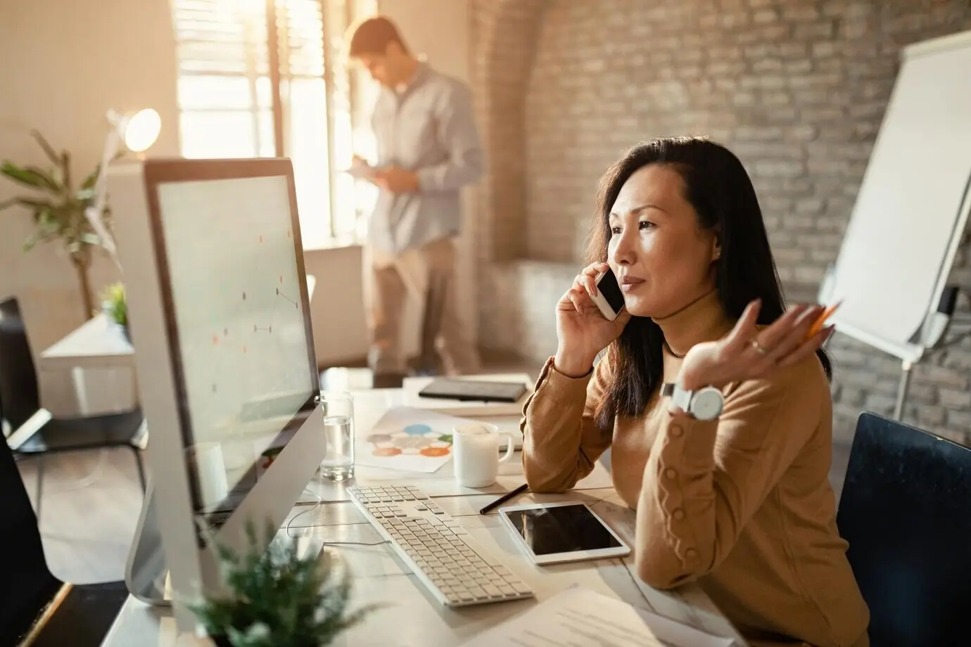 An Asian businesswoman reviewing charts on a desktop PC while communicating on a cell phone in the office.