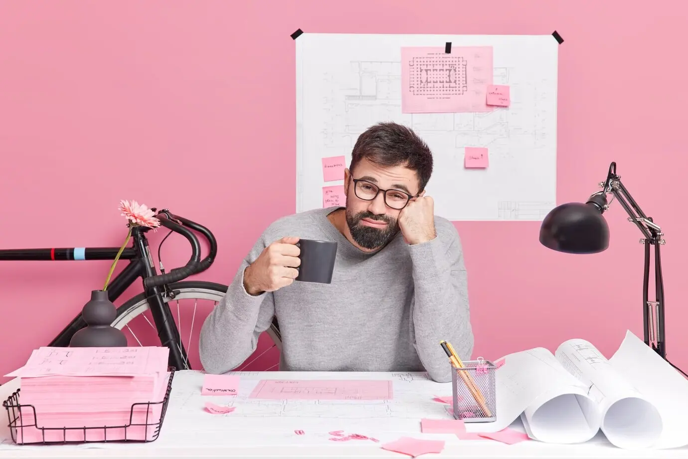 Engineering, construction, and architecture concept. A tired male office worker drinks refreshing coffee and works all night on an urgent task with a deadline to finish the work, posing at a desktop in a coworking space.